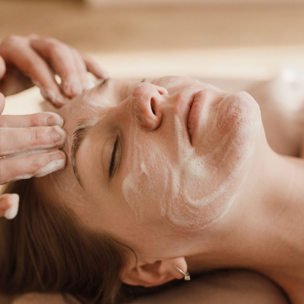 Mujer con los ojos cerrados mientras recibe un tratamiento facial, con las manos de la terapeuta en la frente y la sien
